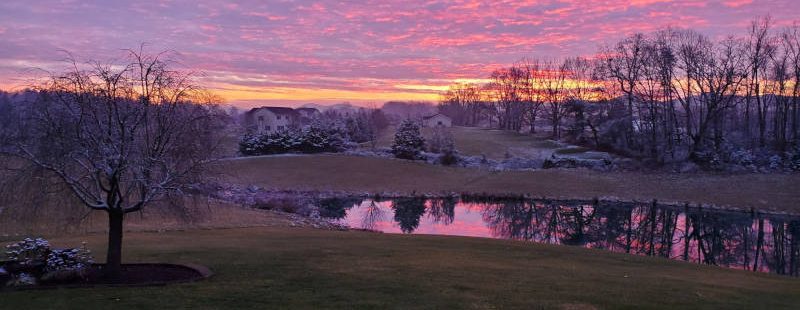A beautiful pink sunset behind a lightly frosted landscape of bare trees and rolling hills beyond a small lake